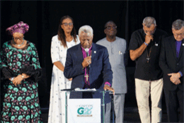 Six people pray with closed eyes behind an African Archbishop at a podium